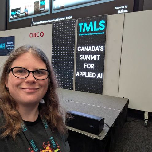 Meagen Voss smiling for a selfie in front of a black and blue sign that says Toronto machine learning summit.