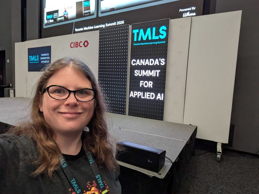 Meagen Voss smiling for a selfie in front of a black and blue sign that says Toronto machine learning summit.