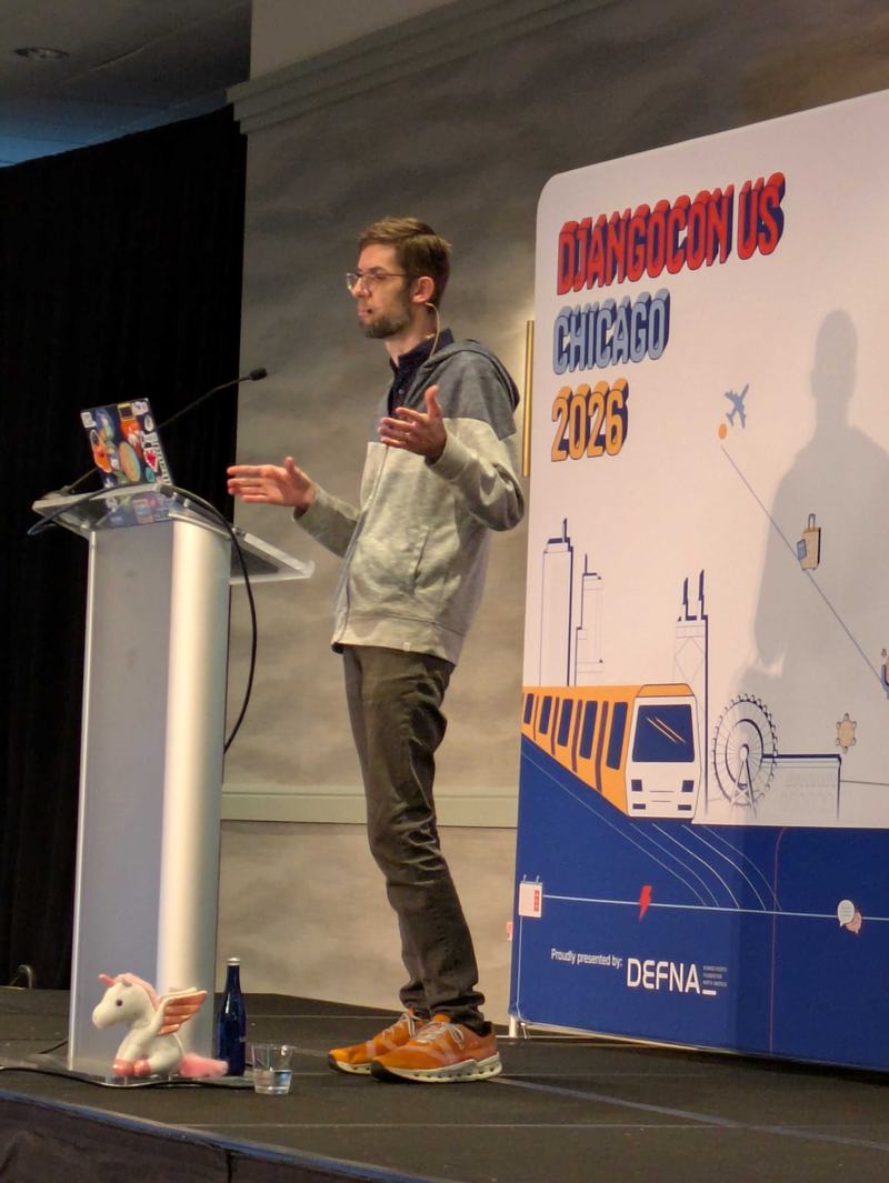 Scott Cranfill speaking on the main stage at DjangoConUS 2026. You can see a pink winged pony close to his feet by the lectern.