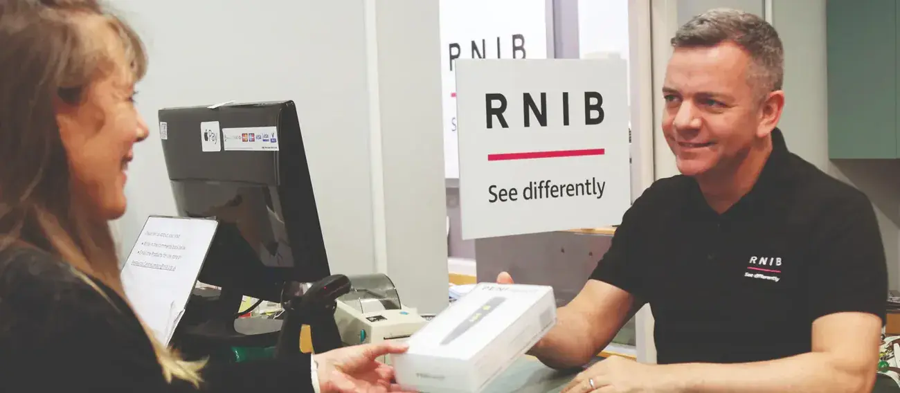 A smiling man with a short brown hair cut and pale skin hands a box to a woman from across a desk. There is a sign that says RNIB in the background.