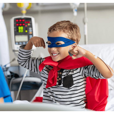 A little boy sitting in a hospital bed wearing a superhero cape and holding his arms up in the air.