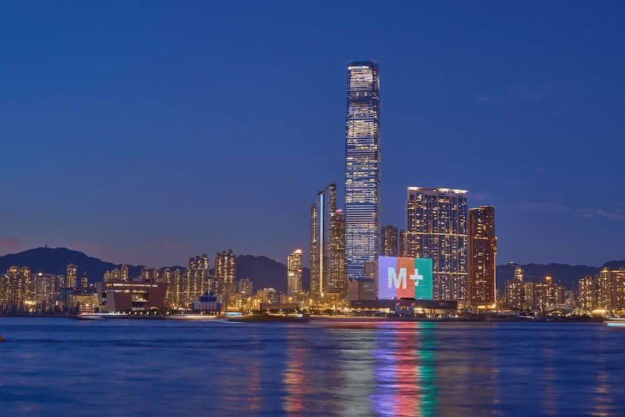 M+Museum building against the Hong Kong skyline at dusk, with the sea in the foreground. The building has a giant "M+" displayed on it, in white with purple - red - turquoise background