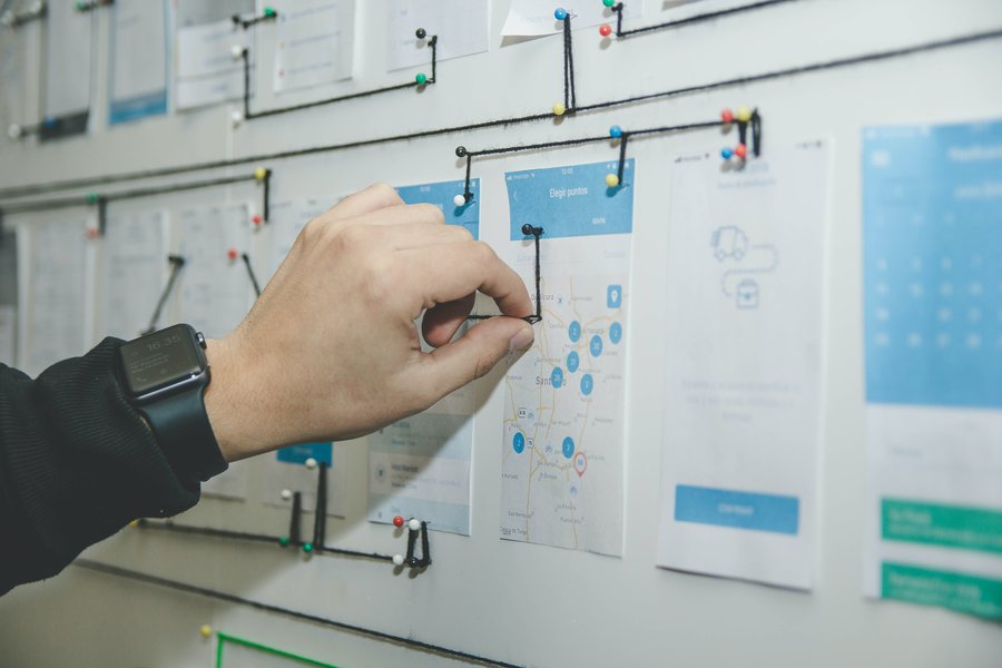 In this image, there is a person organising papers on a whiteboard and pinning pieces of thread from paper to paper like they are organising a workflow.